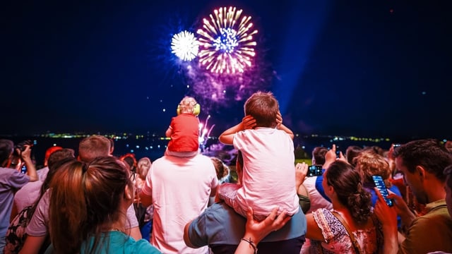 Ein Kind sitzt auf den Schultern eines Mannes und hält sich die Ohren zu. Im Hintergrund ist ein Feuerwerk zu sehen.