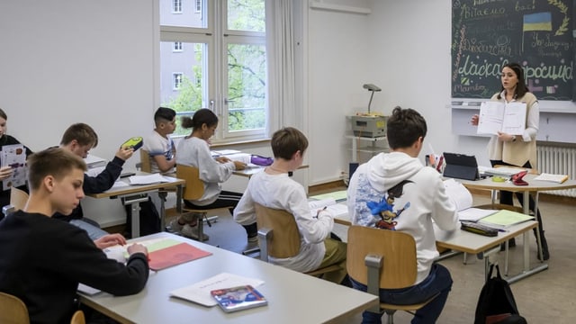 Foto einer Schulklasse auf Bänken, Rückenansicht. Vorne an der Wandtafel steht junge Lehrerin mit offenem Buch.