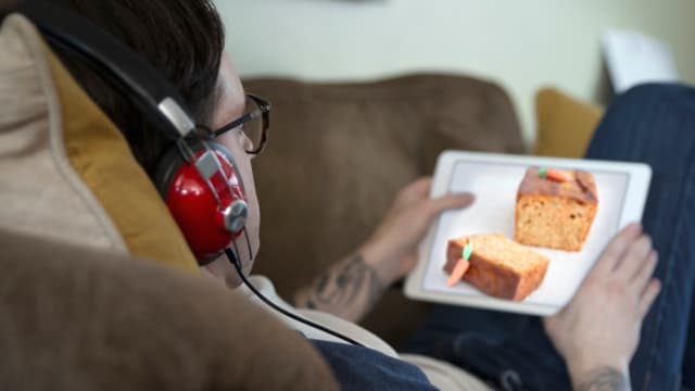 Ein Mann liegt auf einem Sofa und schaut auf ein Tablet, auf dem ein Kuchen zu sehen ist.