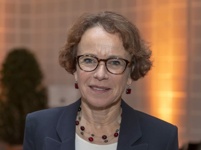 Frau mit kurzen braunen Haare und Brille, dunkelblauen Blazer, weissem T-shirt und rotem Steinschmuck.