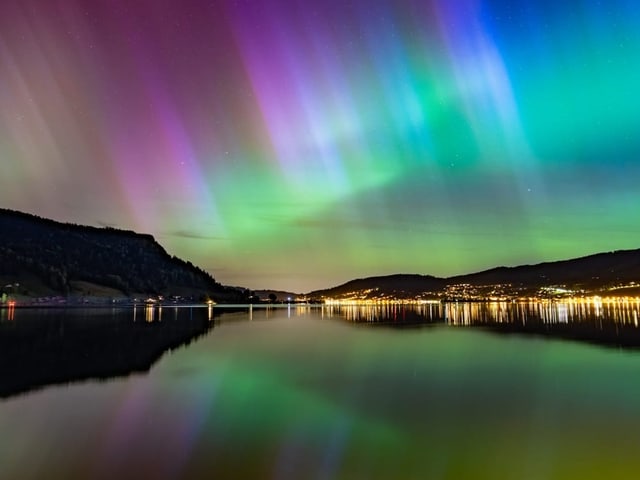 Nordlichter über einem See bei Nacht mit beleuchteter Stadt im Hintergrund.