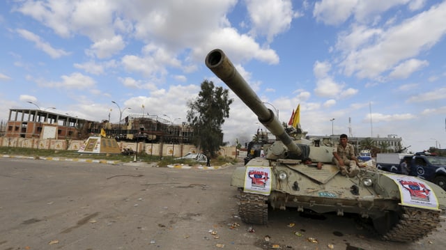 Ein Panzer steht auf einer Strasse, darauf sitzen Soldaten, im Hintergrund zerstörte Häuser.