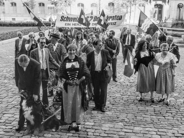 Menschenmenge mit Schweizer Fahnen und traditioneller Kleidung auf Kopfsteinpflaster.