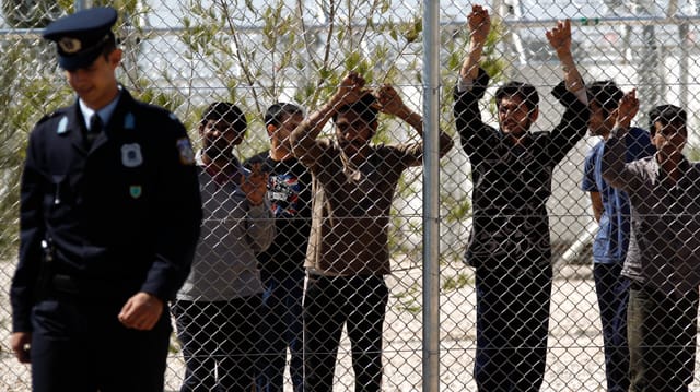 Ein Polizist geht an einem Zaun eines Flüchtlingscamps in Athen vorbei, dahinter sechs Männer.