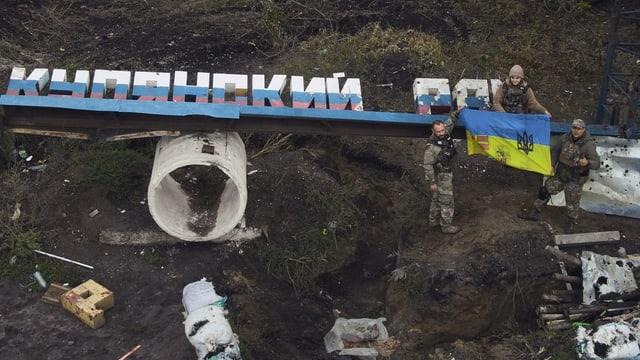 Zwei Männer und eine Frau, die eine ukrainische Fahne an einem zerstörten Schild anbringen.