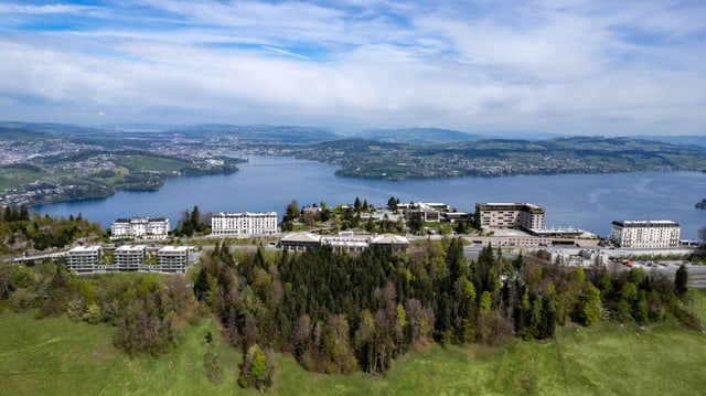 Luftaufnahme von Gebäuden am Seeufer mit grüner Landschaft und bewaldeten Hügeln.