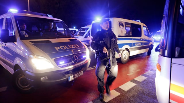 Polizeifahrzeuge stehen mit Blaulicht herum, ein bewaffneter Polizist davor, Szene bei Nacht.