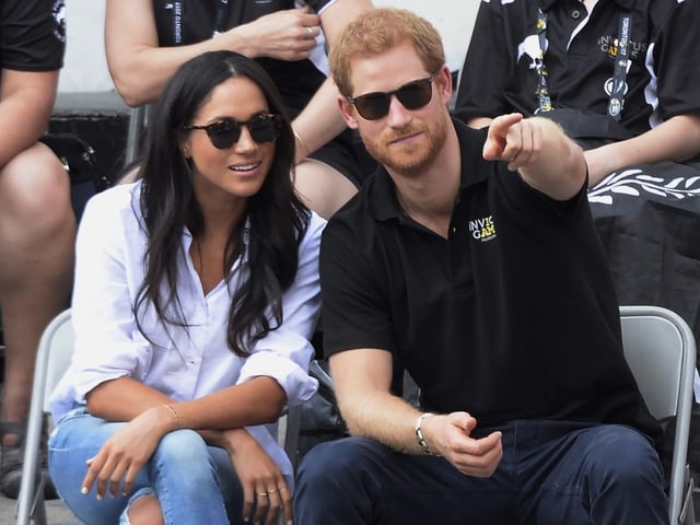 Harry und Meghan sitzen nebeneinander, Harry zeigt Meghan mit dem Finger etwas.