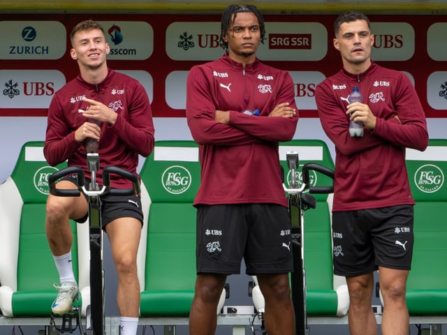 Drei Fussballspieler in Trainingsanzügen mit Sponsoren im Hintergrund.