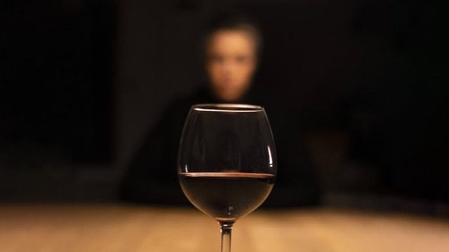 vorne im Fokus. Ein Glas Rotwein. Hinten vor schwarzem Hintergrund ganz verschwommen ein Gesicht.