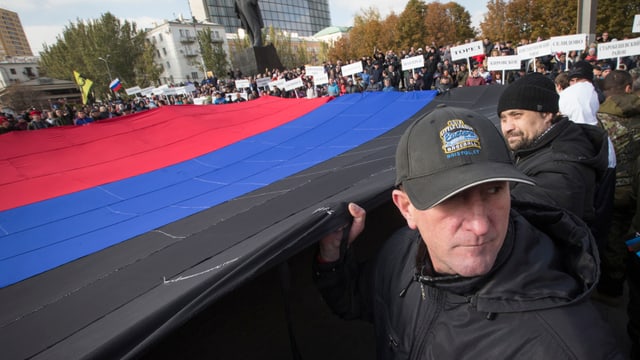 Eine riesige, rot-blau-schwarze Flagge wird von Dutzenden Menschen auf einem Platz gehalten, im Hintergrund eine Lenin-Statue.