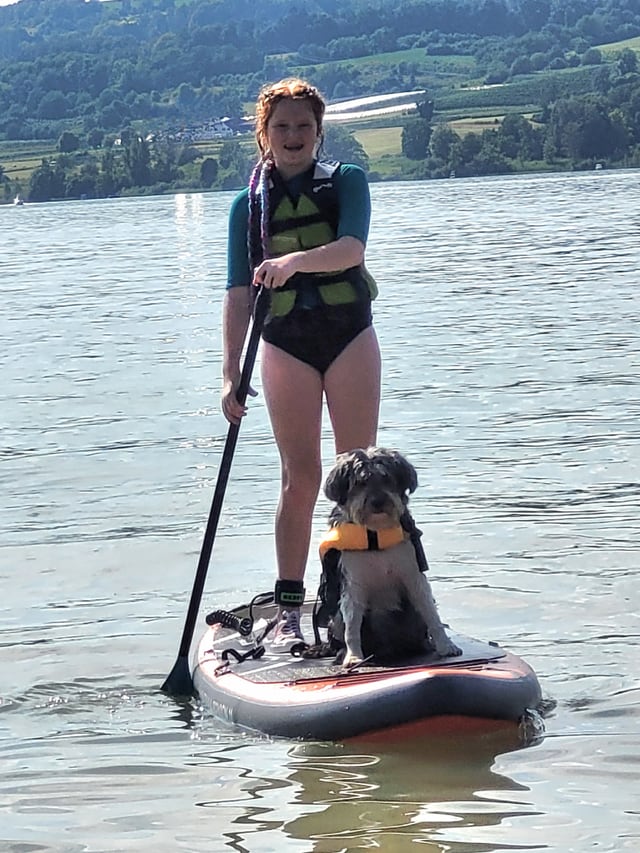 Ein Mädchen steht auf einem Stand Up Paddle Board. Ein Hund sitzt vor ihr auch darauf. 