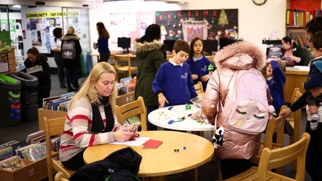 Menschen und Kinder stehen in einer Bibliothek um Tische. Die Kinder spielen mit Spielzeug. Frau liest am Handy.