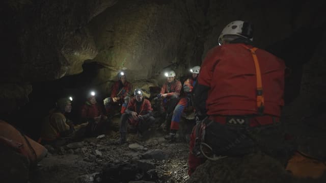 Forschende sitzen in Schutzkleidung in Höhle. Helm mit Lampen auf dem Kopf