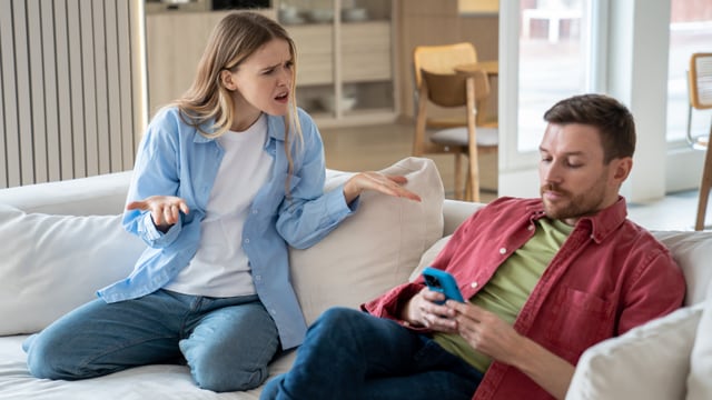 Eine Frau beschimpft einen Mann, der mit dem Smartphone in der Hand neben ihr auf dem Sofa sitzt und sie nicht beachtet.