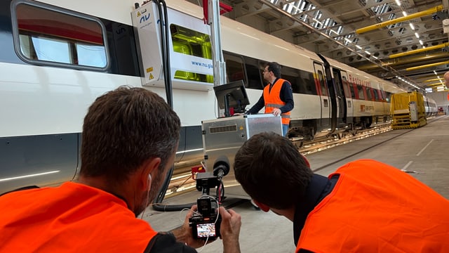 vorne zwei Männer in orangen Westen mit Kamera, hinten Mann mit Metallkasten an Zugscheibe. Langer SBB-Zug.