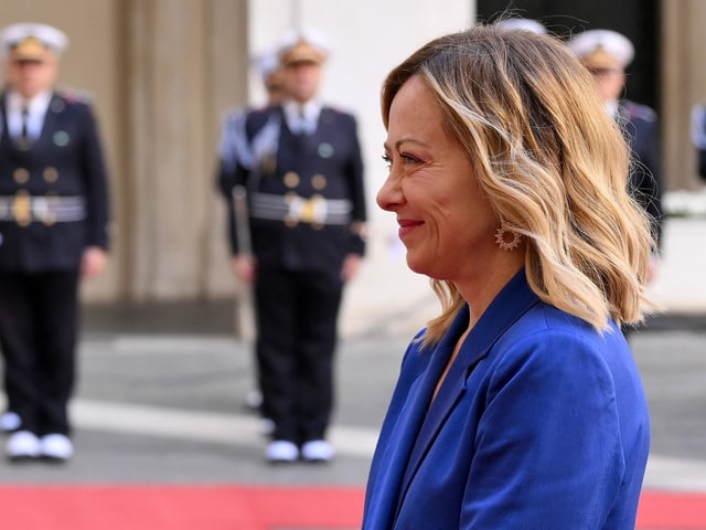 Frau im blauen Blazer, Militärpersonen im Hintergrund.