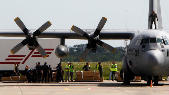 Gestapelte Särge vor einem Flugzeug.