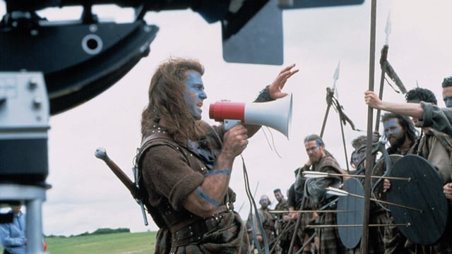 A man with long hair, a kilt and a megaphone in hand, stands in front of a horde of costumed men and gives instructions.