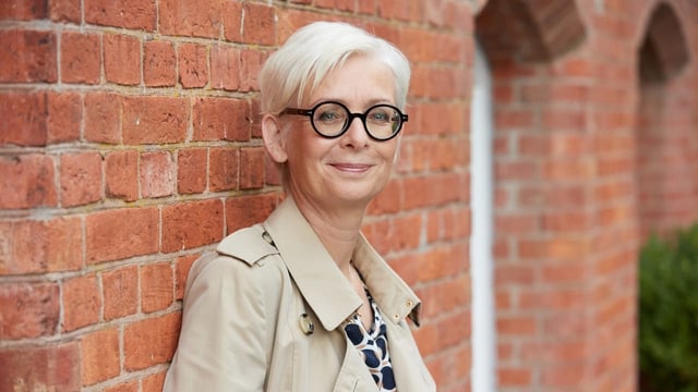 Frau mit hellen Haaren und Brille vor einer Backsteinwand.