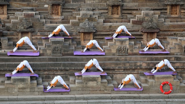 Menschen machen auf einer violetten Yogamatte in weiss-orangen Kleidern einen Sonnengruss. 