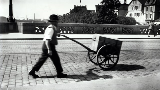 Auf der Schwarz-Weiss-Fotografie schieb ein Mann einen Holzwagen über eine Brücke mit Pflastersteinen.