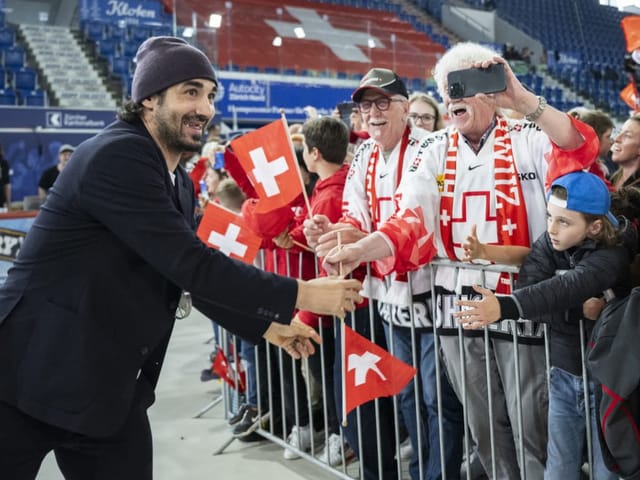 Mann begrüsst Fans mit Schweizer Fahnen in einer Sportarena.