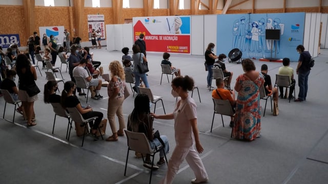 Eltern und Kinder sitzen und stehen in einer Turnhalle und warten auf die Impfung.