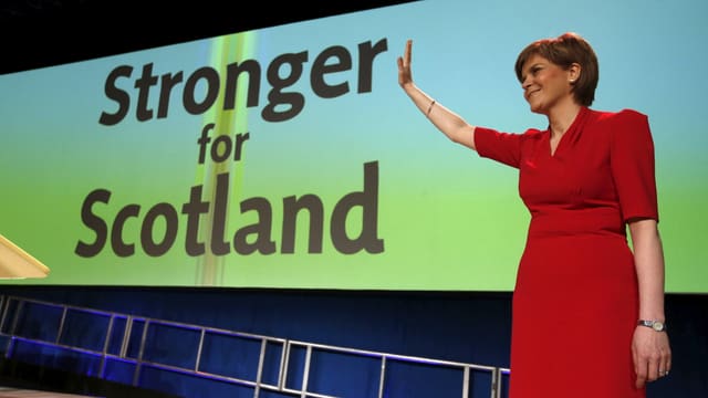 Eine Frau in einem roten Kleid winkt vor einem Plakat mit der Aufschrift «Stronger for Scotland».