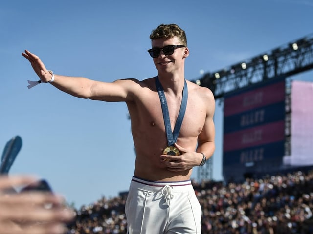 Oberkörperfreier Mann mit Medaille und Sonnenbrille winkt vor einer Menschenmenge.