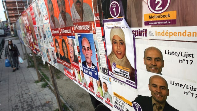 Eine Wand voller Wahlplakate, Eine Frau mit Kopftuch läuft daran vorbei. 