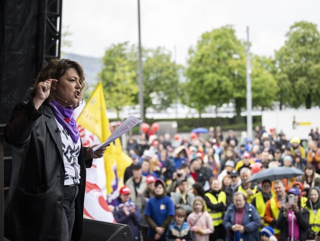 Tamara Funicello steht auf einer Bühne und spricht. Sie hebt einen Finger in die Luft. Im Hintergrund ist Publikum.