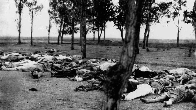 Schwarz-Weiss-Aufnahme: Menschen liegen tot auf einem Feld, auf dem ein paar Bäume wachsen. 