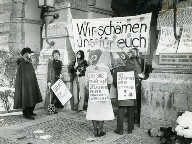 Menschen protestieren mit Schildern und Regenschirmen vor einem steinernen Gebäude.