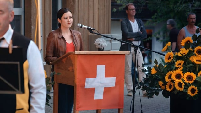 Eine junge Frau hinter einem Rednerpult, das eine Schweizer Flagge ziert.