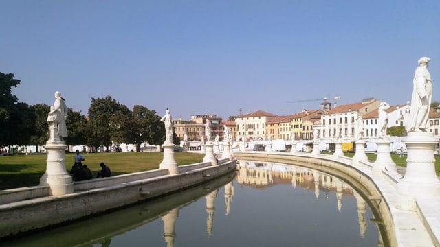 Ein Fluss, gesäumt von alten Gebäuden und weissen Statuen.  Im Hintergrund eine Wiese mit Bäumen.