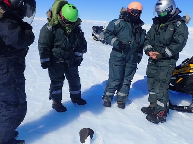 Menschen in dicken Thermoanzügen stehen im Halbkreis um einen Meteoriten im Schnee