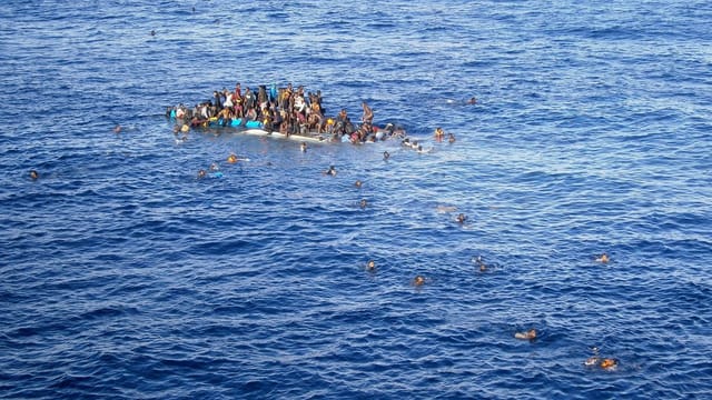 Ein sinkendes Boot, Dutzende Menschen darauf, einige schwimmen im Meer.