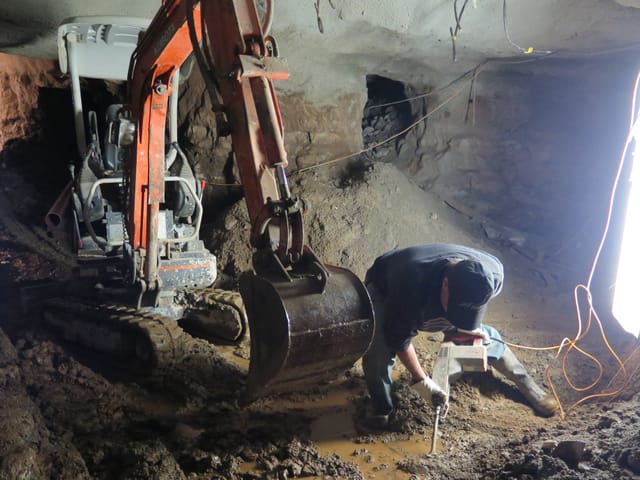 Bagger in einem Stollen. Davor ein Mann mit einer Bohrmaschine.