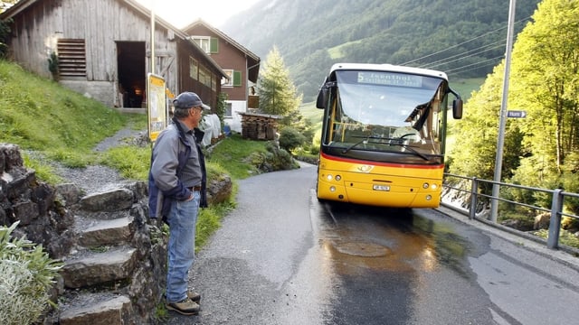 Ein Postauto nähert sich einer Haltestelle, an der ein Mann wartet.