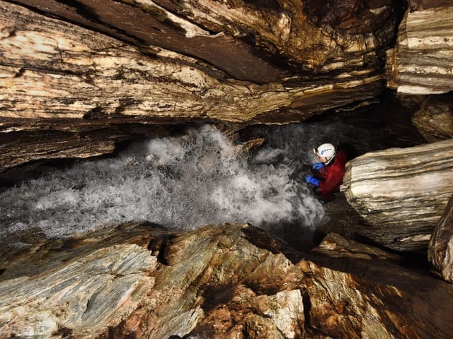 Person mit Helm klettert neben rauschendem Wasser in einer Höhle.