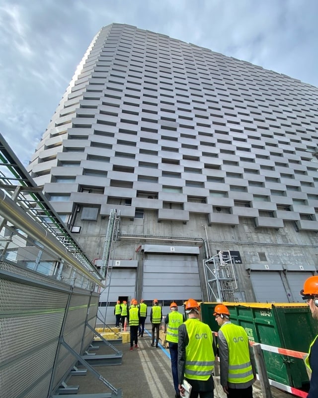 Menschen in Sicherheitswesten und Helmen vor einem Hochhaus.