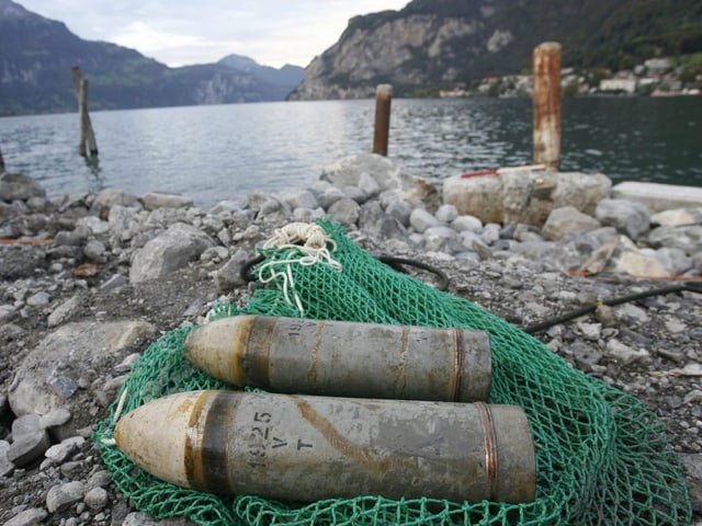 Zwei Artilleriegeschosse auf Felsstrand vor Bergsee.