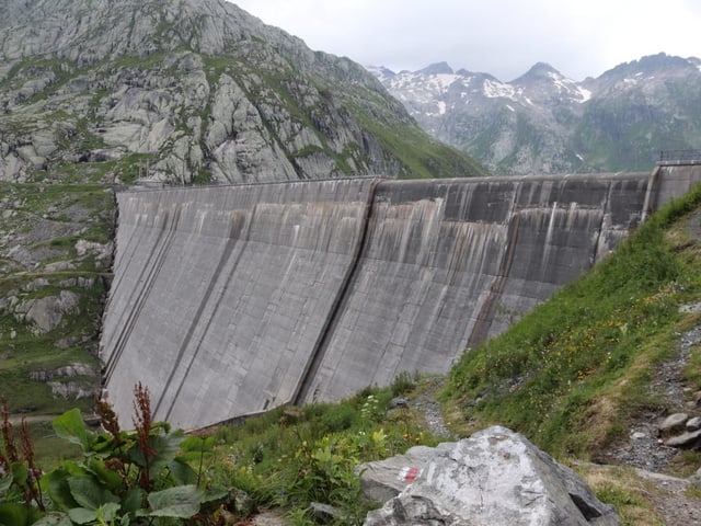 Mächtige graue Staumauer mit Bergen im Hintergrund.