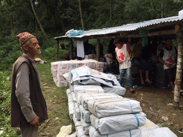 Eingepackte Werkzeugkisten auf einem Stapel. Ein älterer Nepalese steht davor, im Hintergrund Mitarbeiter des Roten Kreuzes.