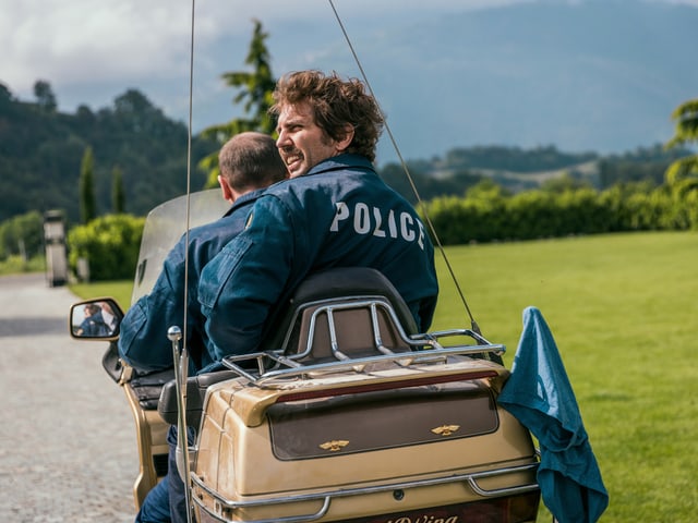 Zwei Polizisten auf einem Motorrad in einer ländlichen Gegend.