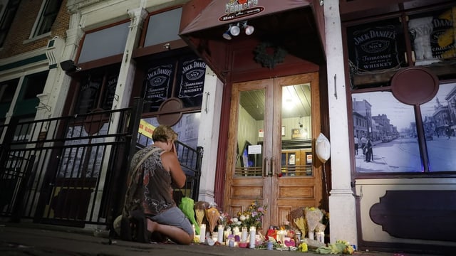 Frau kniet vor dem Eingang einer Bar. Am Boden viele Blumen und Gedenktafeln für die Toten.