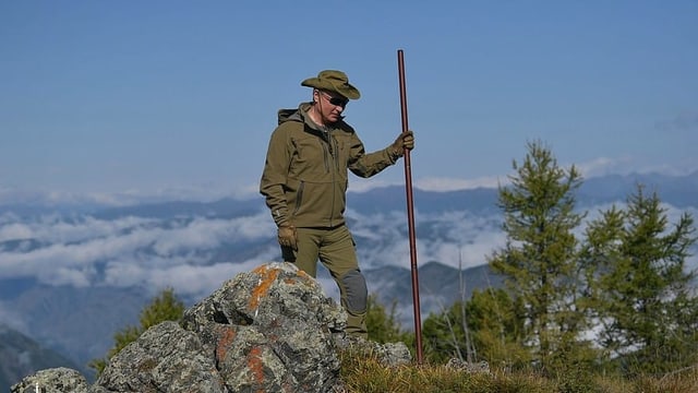 Putin in grüner Outdoorkleidung mit Stock vor blauem Himmel.