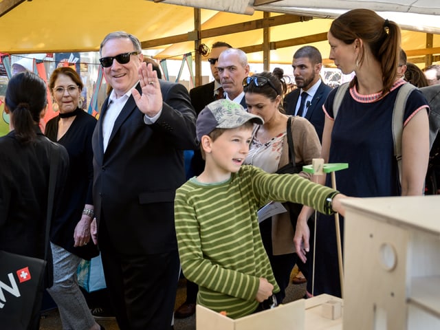 Mann läuft durch Marktstände und grüsst mit der Hand. Ein Kinder steht davor.