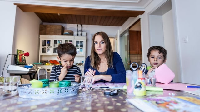 Eine Mutter sitzt am Wohnzimmertisch mit zwei Kindern.